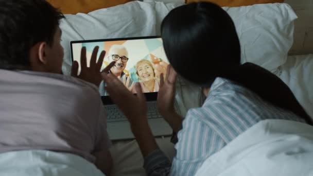 Νεαρό ζευγάρι μιλάει με ηλικιωμένους που κάνουν online βιντεοκλήση με φορητό υπολογιστή στο κρεβάτι — Αρχείο Βίντεο