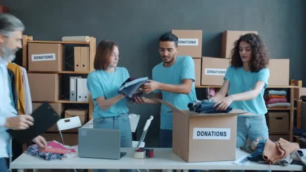 一组志愿者把衣服装在纸板箱里 而慈善公司的经理则在繁忙的办公室的剪贴板上写着 — 图库视频影像