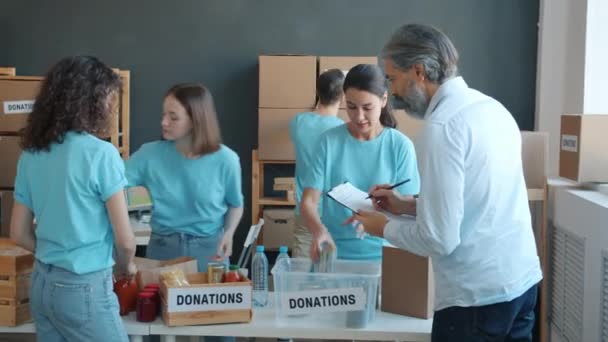 一队慈善工作者把食物装在集装箱里 准备捐给慈善机构 为办公室里的穷人提供帮助 管理员检查剪贴板上的文件 — 图库视频影像