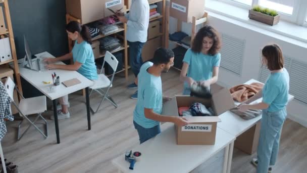 志愿者们一组一组一组准备捐赠服装的盒子 在办公室里一起工作 而成熟的男性经理则在剪贴板上检查名单 — 图库视频影像