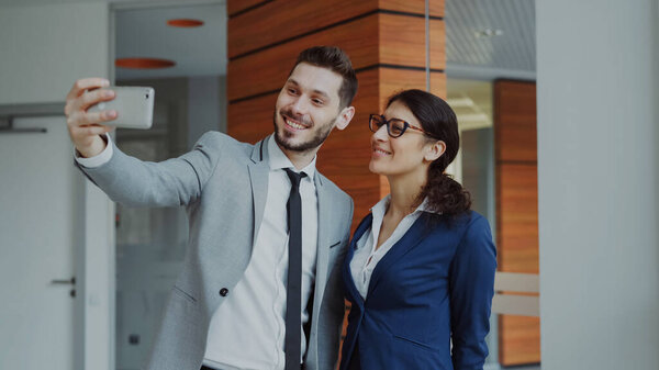 Happy businessman and his female colleague taking a selfie on smarphone camera for social media in modern office indoors