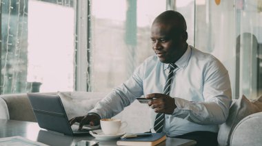 İyimser Afrikalı-Amerikalı iş adamı resmi onun kredi kartı ve dizüstü şık kafede öğle breaak sırasında kullanarak ödeme online fatura giysiler. O mutlulukla gülümser ve onun yumruk el yükseltir.