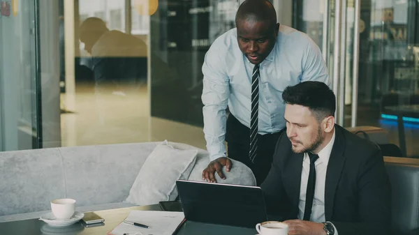 两个商业伙伴积极讨论他们的创业项目 坐在时髦的咖啡馆 胡子的白种商人坐在谈论非洲裔美国同事 而看膝上型计算机 — 图库照片