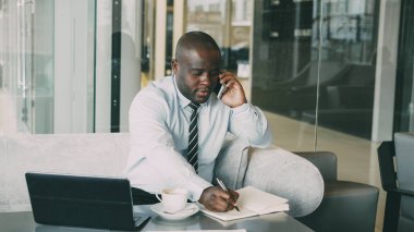 Smartphone üzerinde konuşma ve onun masada onun not defteri dizüstü bilgisayar ve kahve kafede iş bilgileri yazma Afrikalı-Amerikalı işadamı portresi. Çok gesticulates.