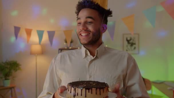 パーティーの帽子保持ケーキで陽気な若い男のスローモーションの肖像画はその後 装飾された部屋で顔でそれを粉砕します 誕生日とお祝いのコンセプト — ストック動画