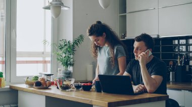 ciddi çift mutfak olarak, kadın salata kesme, sessiz duruyor, adam üstünde laptop çalışma masasında oturuyor ve telefonda konuşurken, o rahatsız ve kızgın.