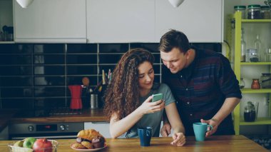 genç çekici kıvırcık mutlu beyaz kadın modern mutfak kapalı manifatura biri smartphone ile oturuyor, kocası geliyor ve onun telefonundaki komik bir şey gösterir