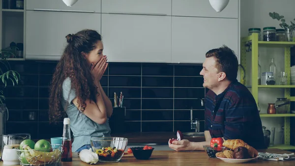 若い男は 彼女の自宅の台所で料理をしながら彼のガール フレンドに提案を行います 彼はキス彼女の指にリングを入れてください — ストック写真