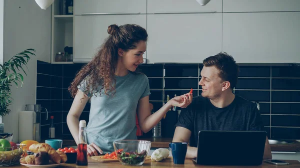 赤唐辛子を試して夫を与えながら テーブルの女性の料理で陽気なカップル 笑顔男座っているラップトップ上で作業 — ストック写真