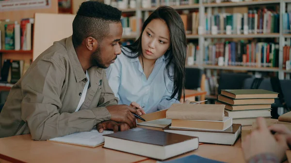 一緒にクラスメートと屋内で大学図書館のテーブルに座って試験の準備作業若いアフリカ系アメリカ人とアジアの大学生 — ストック写真