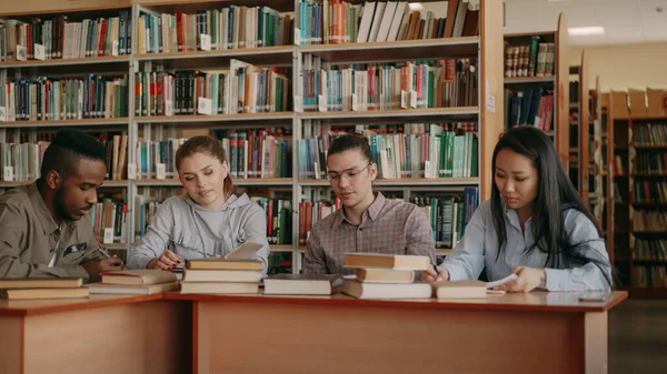 Multi ethnic group of students chatting and preparing for examination while sitting at the table in university library indoors