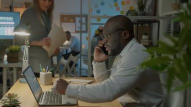 Genç Afrikalı Amerikalı iş adamı cep telefonuyla konuşuyor. Kapalı bir ofiste, geceleri çalışıyor. İletişim ve işgal kavramı.