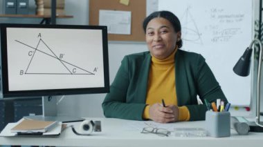Neşeli Afrikalı Amerikalı öğretmen matematiği açıklarken bilgisayar ekranını işaret ediyor ve uzaktaki derste konuşuyor. Kameraya bakan genç bir kadın el sallayarak gülümsüyor..