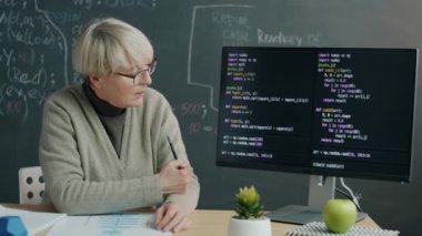 Programlama öğretmeni bilgisayar ekranına işaret eden yazılım kodlamasını açıklıyor ve kurye video dersi sırasında konuşuyor. Uzaktan eğitim ve bilişim sınıfı kavramı.