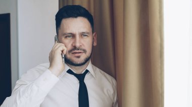 Beyaz gömlek ve kravat cep telefonu ayakta otel odasında pencere yakınındaki konuşurken kendinden emin sakallı işadamı