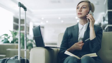 Cep telefonu konuşurken, Not Defteri'nde yazma ve dizüstü bilgisayar gülümseyen kullanarak otel lobisinde koltukta oturan sarışın meşgul iş kadını. İş, seyahat ve insanlar kavramı