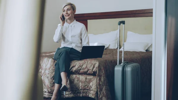 白いシャツのラップトップを使用して ホテルの部屋でベッドの上に座って携帯電話で話している笑顔金髪実業家のパン ビジネス の概念 — ストック写真