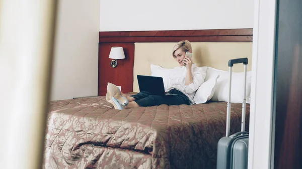 白いシャツのラップトップを使用して 携帯電話で話しながらホテルの部屋でベッドに横になっている笑顔金髪実業家 ビジネス の概念 — ストック写真