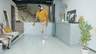 Afrika kökenli Amerikalı adamın mutfakta yerleri yıkaması ve dans etmesi ve kulaklıkla müzik dinlemesi. Ev işleri ve eğlence konsepti.