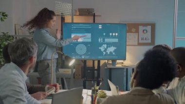 Karanlık ofisteki çeşitli meslektaşlarına sunum yapan genç iş kadını dijital ekrana işaret ediyor ve konuşuyor. İşbirliği ve başlangıç konsepti.