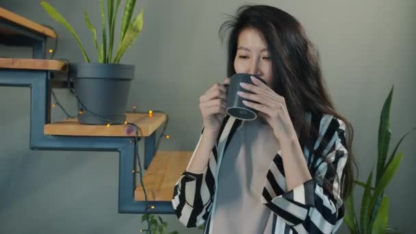 現代のアパートでコーヒーを飲む幸せなアジアの女性は一人で朝を楽しんで笑顔 肯定的な感情と近代的な人々の概念 — ストック動画