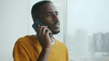 Mutlu genç adam cep telefonuyla konuşuyor. Büyük pencerenin yanında duruyor. Gülümsüyor ve sohbet ediyor. Gençlik ve iletişim kavramı.
