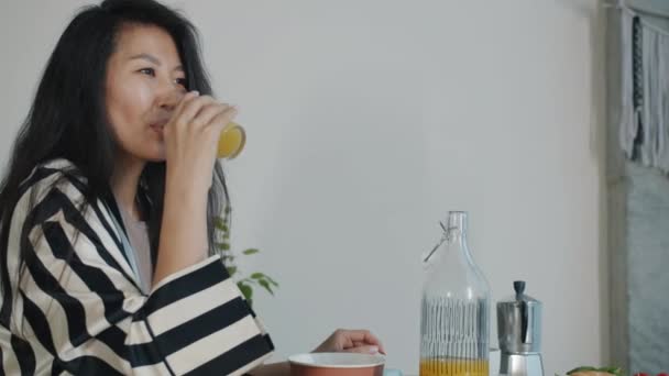 愉快的亚洲女人在家里吃早餐时喝新鲜的橙汁 微笑着喝着喝着 人与健康生活方式概念 — 图库视频影像