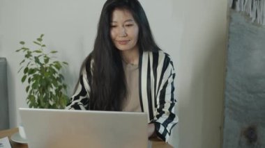 Evde, mutfakta çalışan dizüstü bilgisayarı kullanan başarılı genç Asyalı bayan. Modern teknoloji ve uzak çalışma konsepti.