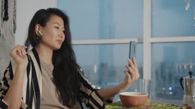 Mutlu Asyalı kadın evde yemek yerken çevrimiçi video görüşmesinden keyif alıyor ve gülüyor. İnternet iletişimi ve gençlik yaşam tarzı kavramı.