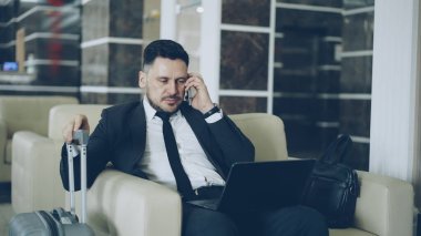 Cep telefonu konuşurken ve lüks Otel Bagaj çanta ona yakın koltukta otururken dizüstü bilgisayar kullanan emin çekici iş adamı yukarı eğimli. Seyahat, iş ve insan kavramı