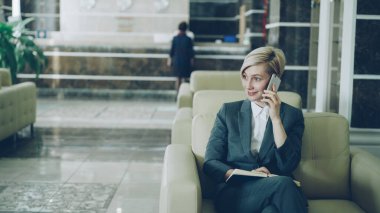 Cep telefonu konuşurken, Not Defteri'nde yazma ve dizüstü bilgisayar gülümseyen kullanarak otel lobisinde koltukta oturan sarışın meşgul işkadını kadeh kaydırmak. İş, seyahat ve insanlar kavramı