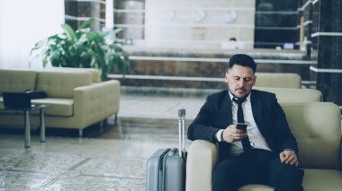 Konsantre işadamı koltuk lüks Otel Bagaj ona yakın oturan smartphone kullanarak kadeh kaydırmak