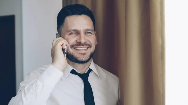 自信微笑的商人在白色衬衣和领带谈话在移动电话站立在窗口附近在旅馆房间里 — 图库照片