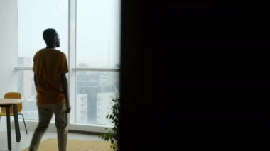 Genç Afro-Amerikan bir adam, günlük giysiler içinde mutfaktaki pencereye doğru yürüyor ve tek başına güzel manzarayı seyrediyor. İnsanlar ve yaşam tarzı kavramı.