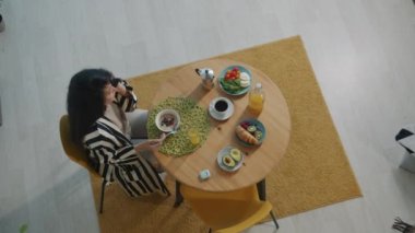 Neşeli Asyalı bir kadının kahvaltı edişinin ve cep telefonuyla sohbet edişinin ve evde gülüşünün en güzel görüntüsü. Pozitif duygular ve iletişim konsepti.