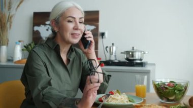 Mutfak masasında cep telefonuyla konuşan neşeli bir bayan ve evde lezzetli bir akşam yemeği. İletişim ve ev hayatı kavramı.