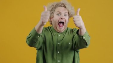 Neşeli genç adam mutluluktan çığlık atıyor ve sarı arka planda duran kameraya bakarak el hareketi yapıyor. Heyecan ve neşe kavramı.