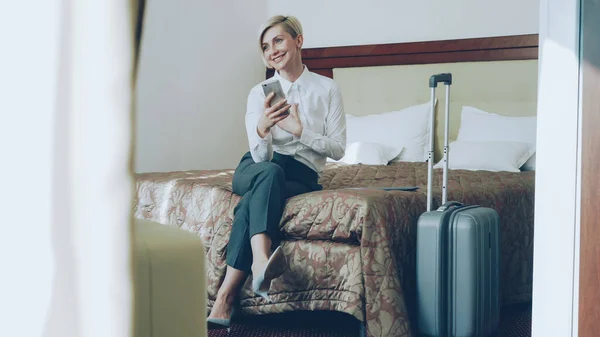 ホテルの部屋でベッドの上に座っている Smrtphone カメラを使用してビデオ チャット経由で家族と話して幸せな陽気な実業家 ビジネス の概念 — ストック写真