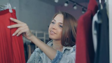 Çekici kız butik seçerek giysilerle yakın çekim shot. O moda üstleri, Kazaklar ve etek onlara dikkatle yüzündeki gülümseme ile bakmak askı üzerinde geçiyor.