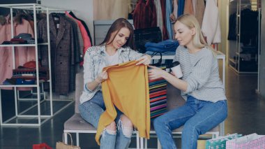 Neşeli kadın müşteri arkadaşı gösterilen sarı jumper birlikte lüks butik otururken satın aldı. Kızlar fiyat ve kalite giyim hakkında heyecanlıyız..