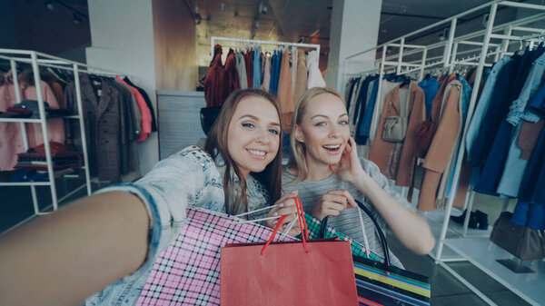 Point of view shot of attractive young ladies making selfie with paper bags in womens clothes boutique. Girls are posing, chatting, and laughing happily. Modern lifestyle concept.