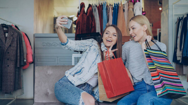 Cute girls are sitting and making selfie with colourful paper bags using smartphone in luxurious womens clothing boutique, then watching pictures together. They are smiling and laughing carelessly.