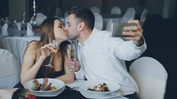 魅力的な夫婦はレストランでロマンティックなディナーをしながらスマート フォンを使用してシャンパン グラスと Selfie を取っています 笑みを浮かべて カメラを見てポーズ — ストック写真