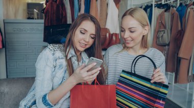 Dikkatsizce sohbet ve güzel giyim butik otururken smartphone kullanarak neşeli genç kadınlar. Onlar gülümseyen ve heyecanla el hareketi. Modern yaşam kavramı.