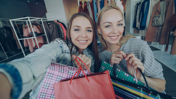 Point of view shot of attractive young ladies making selfie with paper bags in womens clothes boutique. Girls are posing, chatting, and laughing happily. Modern lifestyle concept.