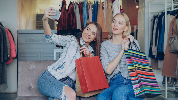 Attractive female friends are using smartphone to make selfie while sitting in womens clothing shop with lots of colourful paper bags. They are smiling, posing, laughing, gesturing.