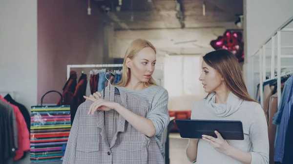 Young business owner is using tablet while standing in her clothing store. Her assistant is coming with garment is hands and asking advice about it. Businesswoman is showing her where to place it.