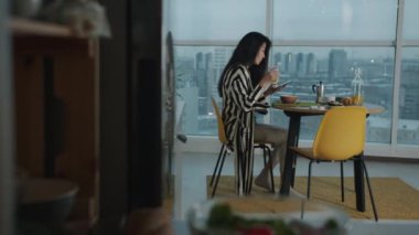 Neşeli Asyalı kadın evde yemek yiyor ve akıllı telefon mesajları atıyor ve mutfakta gülümsüyor. Modern teknoloji ve gençlik yaşam tarzı kavramı.