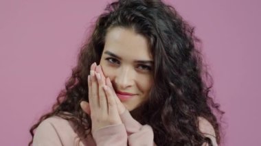 Mutlu genç bir kadının yakın çekim portresi. Yüzü ellere değiyor. Gülümsüyor, pembe arkaplana tapıyor..