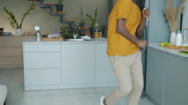 Mutfakta kaşıkla şarkı söyleyen neşeli genç adam evde tek başına eğleniyor. Kablosuz kulaklık takıp müzik dinleyen komik adam..